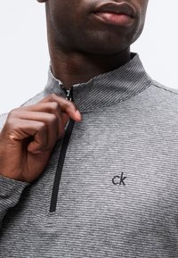 Άνδρας που φορά γκρι ριγωτό φούτερ με φερμουάρ, τραβάει το φερμουάρ κοντά στον λαιμό, με μικρό λογότυπο "ck" στο αριστερό στήθος.