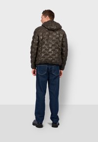 Man wearing dark green quilted hooded jacket, blue jeans, and black shoes standing with back facing camera against plain white background.