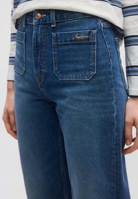 Σκούρα μπλε τζιν με ψηλή μέση, ίσια γραμμή και πορτοκαλί ραφές. Διαθέτουν πίσω τσέπες και ένα κεντημένο λογότυπο μπροστά.