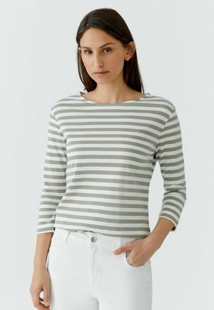 Femme aux longs cheveux foncés portant une chemise à manches longues rayée gris et blanc et un pantalon blanc, debout avec une main dans la poche.