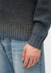 Sivi pleteni pulover z rebrastimi manšetami, ki prehaja iz temnejše v svetlejšo odtenek, kombiniran s svetlo modrimi kavbojkami. Opažena je mehka tekstura.