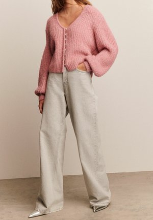 Femme portant un cardigan rose doux boutonné avec des manches ballon, un jean gris clair à jambes larges, et des chaussures argentées pointues, debout avec une main dans la poche.