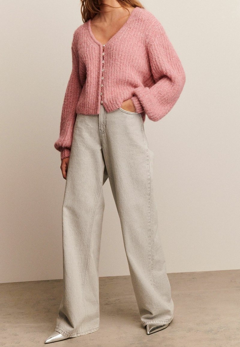 Femme portant un cardigan rose doux boutonné avec des manches ballon, un jean gris clair à jambes larges, et des chaussures argentées pointues, debout avec une main dans la poche.