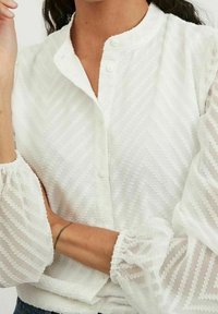 Valkoinen paita, jossa on teksturoitu sahalaitakuviot, nappikiinnitys ja puffihuput, joissa on rypytetyt hihansuut. Suunniteltu keveästä ja hengittävästä kankaasta.