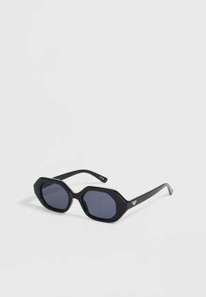 Lunettes de soleil hexagonales noires avec verres foncés et branches fines comportant un petit logo cœur blanc sur le côté.