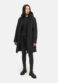 Parka noire avec capuche, fermeture éclair, poches latérales et cordons de serrage réglables, portée sur un haut noir et un jean, associée à des bottes noires.