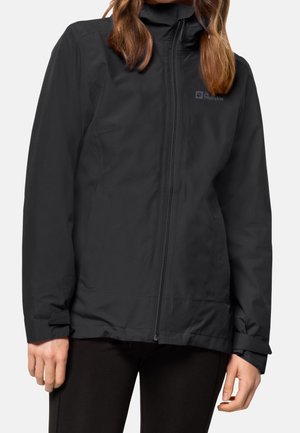 Vrouw draagt een zwart jas met ritssluiting, hoge kraag en verstelbare mouwboorden, staand tegen een effen achtergrond.