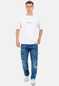 T-shirt blanc à manches courtes avec un logo noir écrit à la main, accompagné d'un jean cargo bleu avec de multiples poches et un motif délavé. Baskets blanches.