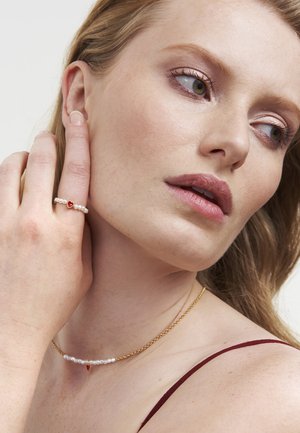 Gouden ketting met kleine witte parels en een rode hartaccent. Eenvoudige ring met een vergelijkbaar ontwerp en een centraal rood hart.