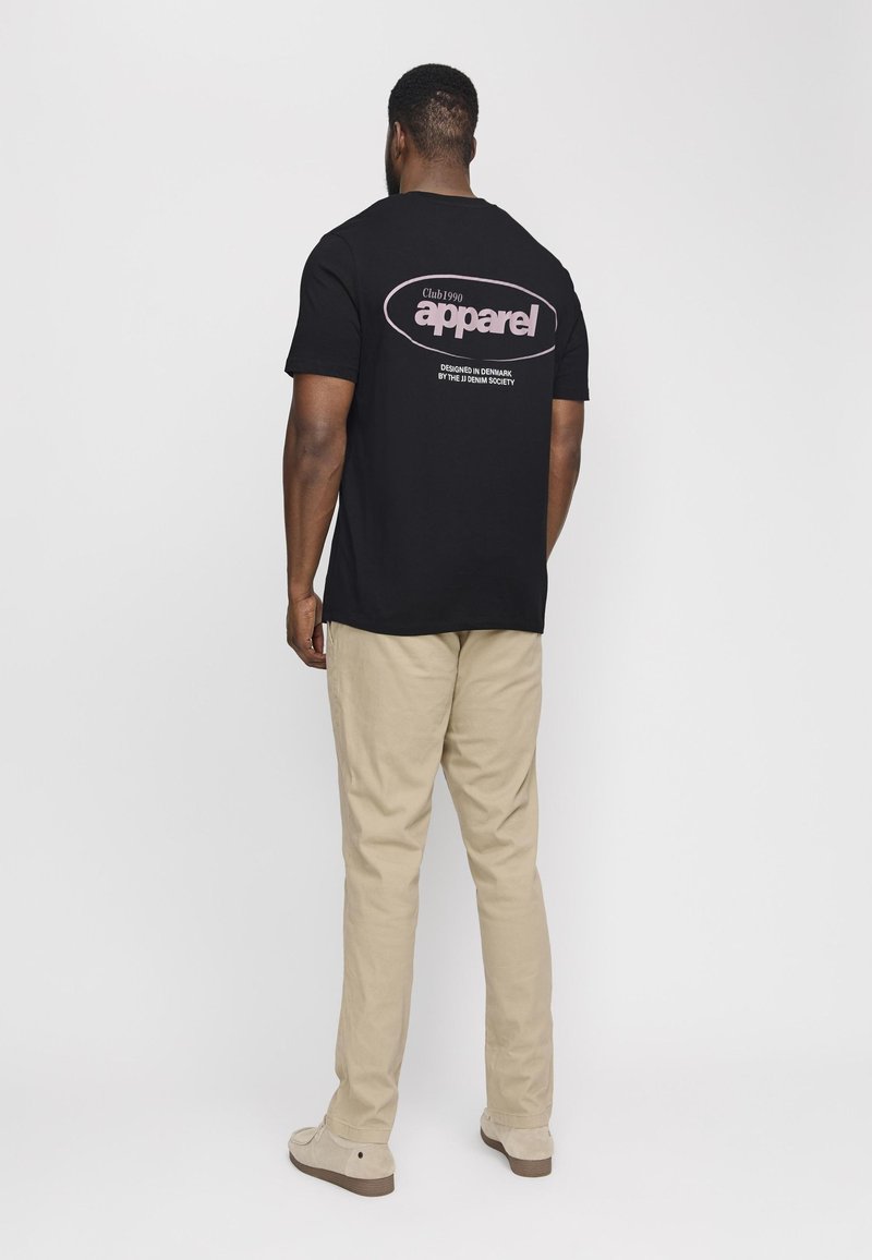 Svart bomullst-skjorte med stor "apparel" grafikk på baksiden. Kombinert med beige bukser og lyse sko. Enkel, avslappet passform.