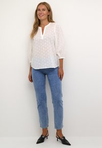 Blusa blanca ligera con patrón texturizado bordado y cuello con muesca. Combinada con jeans de mezclilla azul claro y mules negros de tacón.