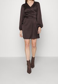Vestido marrón con mangas largas abullonadas, escote en V y cintura con lazo. Se lleva con botas de tobillo marrones que presentan un diseño texturizado.