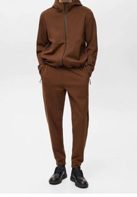 Felpa con zip marrone dotata di grandi tasche, abbinata a pantaloni tapered coordinati. Indossata con sneakers nere. Texture liscia, design moderno.