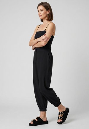 Vrouw staat met gekruiste armen, draagt een zwart mouwloos topje, losse zwarte broek en zwarte instapsandalen met gouden gespen.