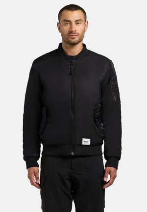 Giacca bomber nera con zip frontale, polsini e vita a costine. Presenta una tasca laterale e un'etichetta con logo sull'orlo inferiore. Texture liscia.