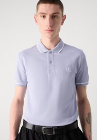 Helllila Polo-Shirt mit klassischem Kragen, zwei Knöpfen und weißen Akzenten an den Ärmelabschlüssen und am Kragen. Dezentes Logo auf der Brust.