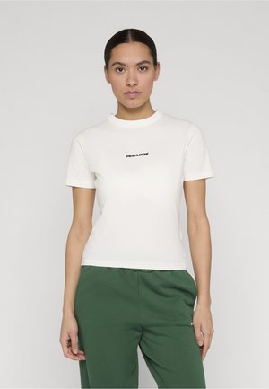 Kvinde med mørkt hår iført en hvid T-shirt med korte ærmer og teksten "PEGADOR", iført grønne bukser, stående mod en ensfarvet lys baggrund.