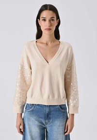 Maglione beige con scollo a V e maniche lunghe, decorato con dettagli in paillettes, dalla texture morbida e orlo a coste. Abbinato a un denim azzurro chiaro.