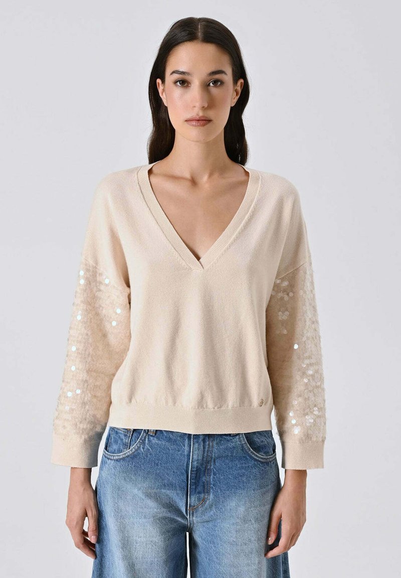 Maglione beige con scollo a V e maniche lunghe, decorato con dettagli in paillettes, dalla texture morbida e orlo a coste. Abbinato a un denim azzurro chiaro.