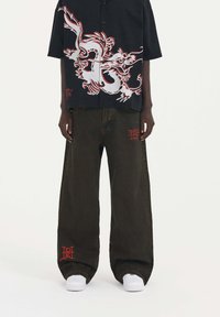 Camisa negra de manga corta con estampado de dragón rojo; pantalones anchos verde oscuro con acentos rojos; combinados con zapatillas blancas.