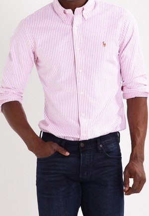 Hombre con camisa de botones de rayas blanco y rosa claro, con las mangas remangadas y vaqueros azul oscuro, una mano en el bolsillo.