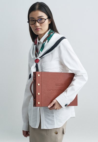 Ung kvinde med langt mørkt hår og briller bærer en hvid skjorte og et mønstret tørklæde, mens hun holder en stor brun Moschino Campionario bog clutch.