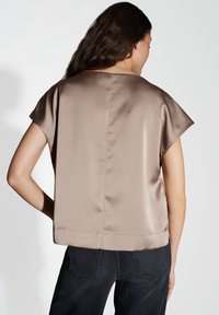 Top corto a maniche corte, di forma boxy, realizzato in un tessuto liscio color taupe. Caratterizzato da un ampio scollo e una cucitura posteriore dritta. Abbinato a pantaloni in denim scuro.
