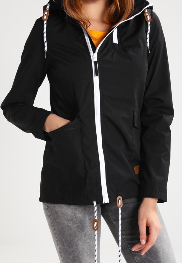 Femme portant une veste noire zippée avec des cordons blancs et des accents marron, associée à un jean gris, main dans la poche de la veste.