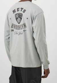 Hellgrau langärmliges T-Shirt mit dem Logo "Nets Brooklyn", NBA-Branding und schwarzen Textdetails auf der Rückseite. Weicher Stoff mit lässiger Passform.