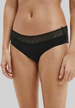 Woman wearing black lace-trimmed Calvin Klein bikini-style underwear standing against a plain light background.