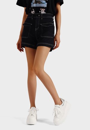 Shorts in denim scuro con tasche frontali, cuciture a contrasto e vita alta, abbinati a sneaker bianche con zeppa e una t-shirt nera con grafica.