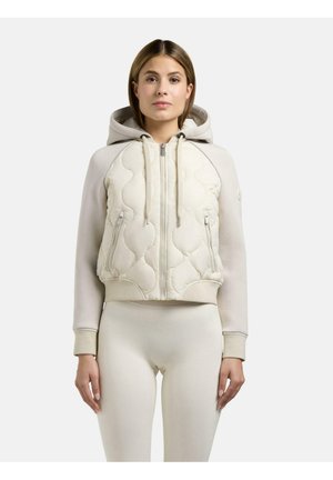 Kvinde iført en cremefarvet, quiltet hættetrøje med lynlås og forlommer samt matchende tætsiddende leggings, stående mod en ensfarvet lys baggrund.