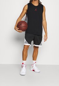 Černý bezrukávový sportovní top s červeným logem, doplněný o šedé šortky s bílým lemem. Bílé a červené basketbalové boty outfit dokončují.