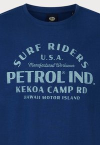 Blauwe katoenen t-shirt met versleten witte tekst "SURF RIDERS U.S.A. PETROL IND. KEKOA CAMP RD HAWAII MOTOR ISLAND." Korte mouwen, ronde hals.