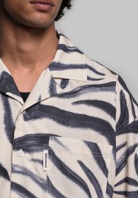Camicia a maniche corte di colore crema con strisce zebra nere; presenta una tasca e chiusura con bottoni. Il tessuto appare liscio e leggero.