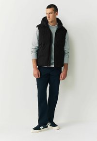 Schwarze Steppweste mit Kapuze über einem grauen Pullover, kombiniert mit navyfarbenen Hosen und navyfarbenen Sneakern mit einem weißen Logo. Minimalistisches Design.