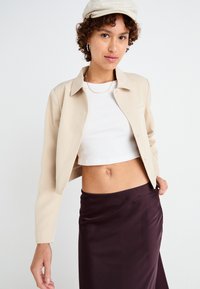 Beež cropped jakk koos nööpide ees, valge ribaroheline tank top ja tumepruun satiinseelik. Mudel kannab beeži mütsi ja rõngas kõrvarõngaid.