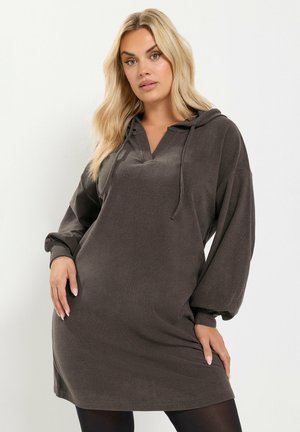Femme blonde portant une robe ample à capuche gris foncé avec de longues manches bouffantes, debout devant un fond clair uni.