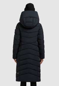 Garšs melns pūkains mētelis ar chevron šuvēm, lielu kapuci un smalku zīmola logotipu apakšējā kreisajā pusē. Gluda tekstila apdare.