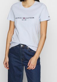 Vaaleansininen puuvillapaita, jossa on tummansinisellä ja punaisella "TOMMY HILFIGER EST. 1985" -teksti. Lyhyet hihat, pyöreä kaula-aukko, rento malli.