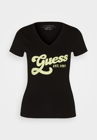 Sort V-hals T-shirt lavet af bomuld; har et stort, lysegrønt "Guess" logo med "EST. 1981" nedenunder, glat tekstur, korte ærmer.