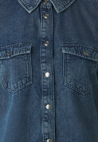 Mørkeblå denimskjorte med knappestolpe foran, to brystlommer og kragehals, som fremhever teksturert stoff og metalliske knapper.