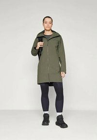 Chaqueta impermeable verde oliva con cremallera en la parte delantera, mangas largas y capucha ajustable, combinada con leggings negros ajustados y zapatillas oscuras.