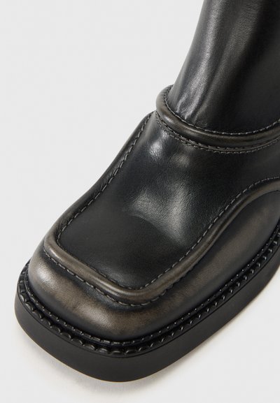 Μαύρο δερμάτινο μποτάκι με ομαλή υφή, τετράγωνη μύτη, ανυψωμένες ραφές κατά μήκος της άκρης και αντί θετη μαύρη ελαστική σόλα.