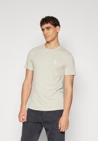 Svetlo béžové tričko s krátkym rukávom vyrobené z mäkkej bavlny, ktoré má minimalistické nášivku na ľavej hrudi a klasický okrúhly výstrih.