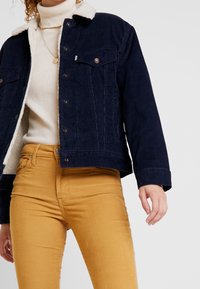 Kvinna iklädd senapsgula kordbyxor, krämfärgad polotröja och marinblå kordjacka med vit fleece-foder.