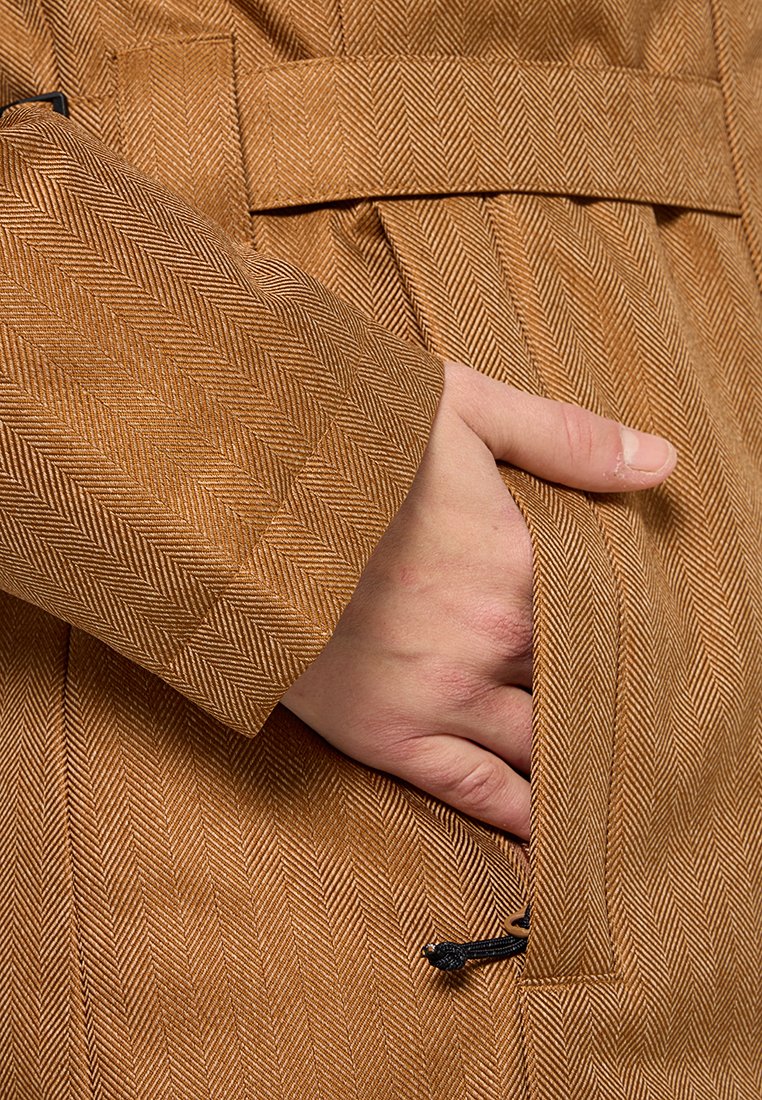 Braune Jacke im Fischgrätenmuster mit strukturierter Oberfläche; verfügt über eine Seitentasche und sichtbare Kordeldetails an der Taille.