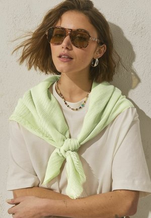 Noor pruunide lühikeste juustega naine, kes kannab kilpkonnamustriga päikeseprille, valget särki, helerohelist kampsunit õlgade ümber seotud ja helmestega kaelakeed.