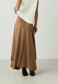 Hnědá sukně s vysokým pasem a asymetrickým lemem, vyrobená z měkkého materiálu; kombinována s bílým topem bez rukávů a černými vysokými botami.