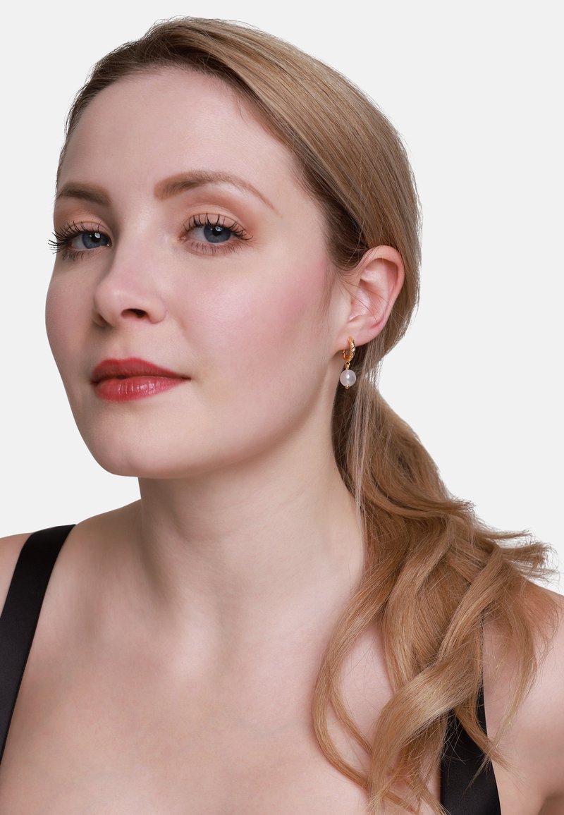 Orecchini a cerchio d'oro con un pendente di pietra chiara. La modella ha i capelli biondi lisci acconciati su un lato, indossa una maglietta nera. Pelle chiara e trucco neutro.
